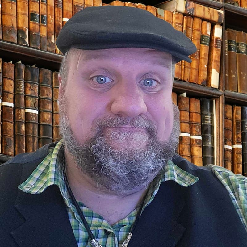A photo of Will Walker in what appears to be a library or study. He looks directly at the camera with a curious expression, wearing a green plaid shirt, black vest, and a dark flat cap, set against a backdrop of floor-to-ceiling antique books.