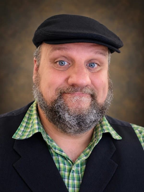 A portrait of Will Walker, a bearded man with blue eyes and a playful, wide-eyed expression. He wears a black flat cap, a green plaid shirt, and a black vest against a warm, mottled brown backdrop.